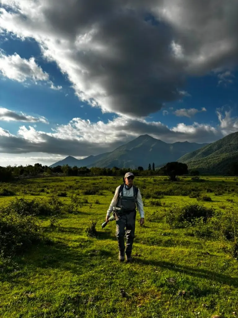 Pescador cruza un prado verde camino al rio, con la cordillera y nubes de tormenta al fondo.