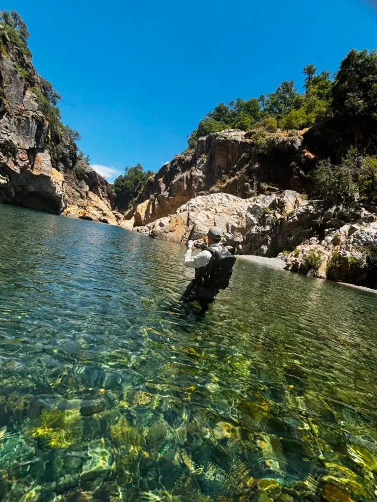 Pescador vadea un canon de aguas claras donde se ve el fondo de piedras.