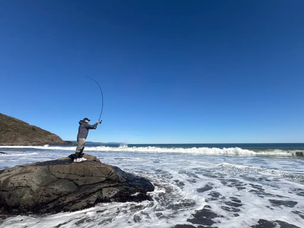 Pescador de pie sobre una roca lanza hacia el mar mientras rompe una ola frente a el.