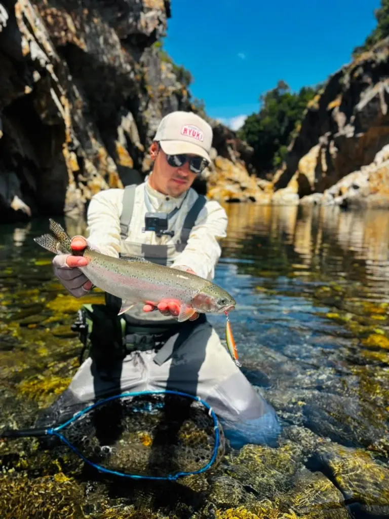 Pescador sostiene una trucha arcoiris sobre el agua, dentro de un canon de paredes de roca.